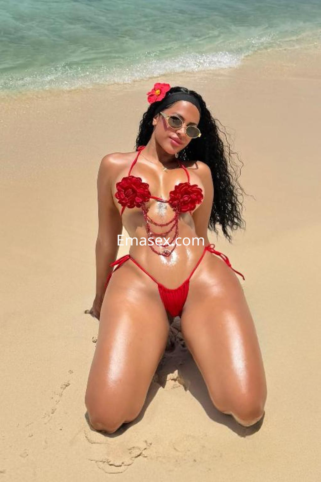La puta en Ibiza luce un bikini rojo adornado con flores, posando sobre la arena junto al mar.