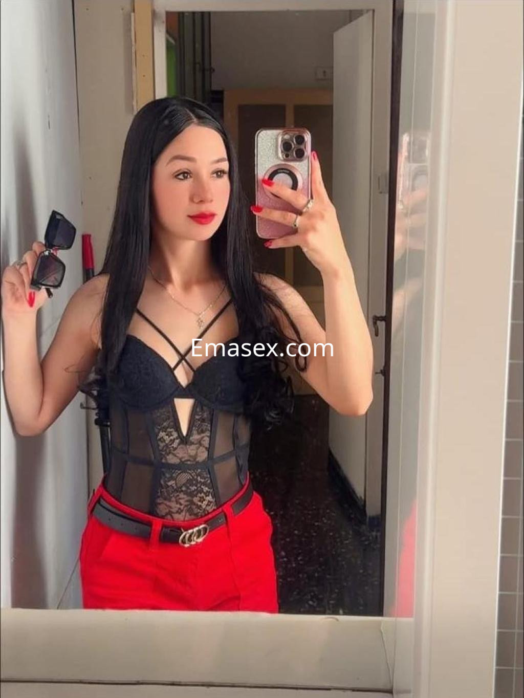 Escort en Pamplona, con cabello largo y lacio, viste un body negro de encaje y pantalones rojos, posando en un espejo.