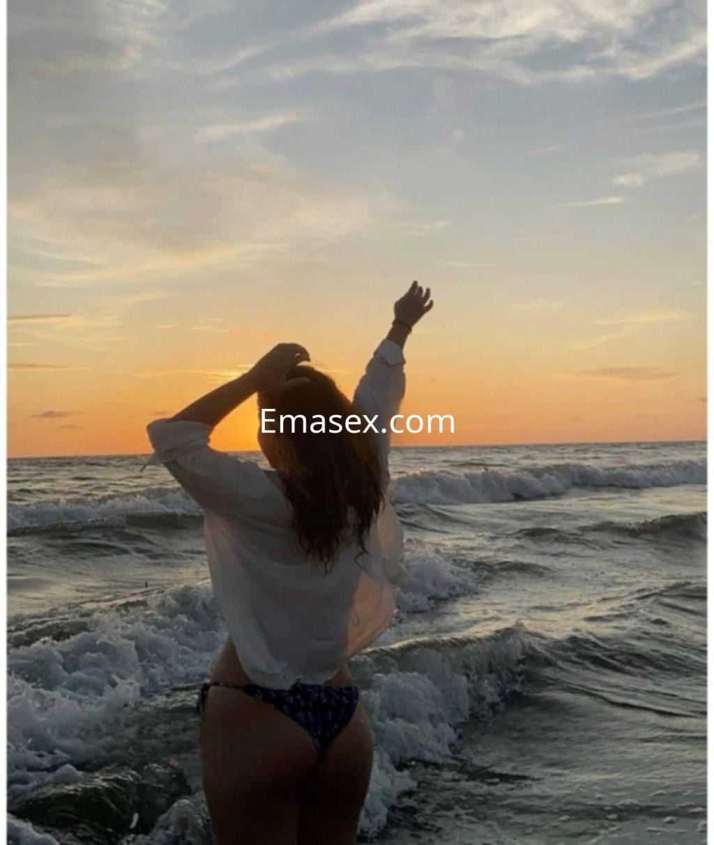 Puta en Zamora de espaldas frente al mar al atardecer, luciendo una blusa blanca y bikini, con el brazo levantado.