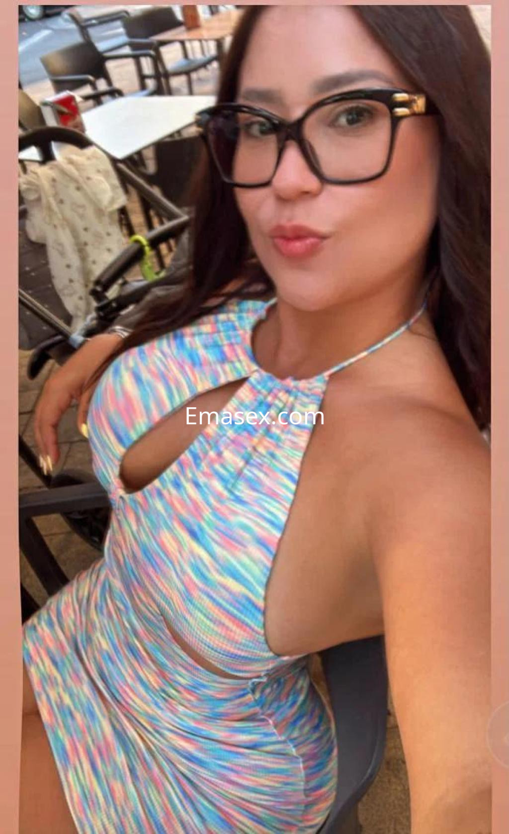 Escort en Sevilla con un vestido de colores pastel, luciendo gafas y posando sonriente en una silla.