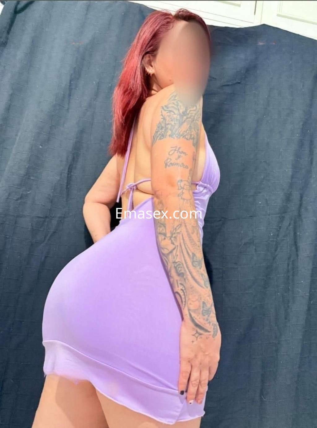 Escort en Sevilla con vestido morado ajustado y tatuajes visibles, posando de lado con el cabello rojo.