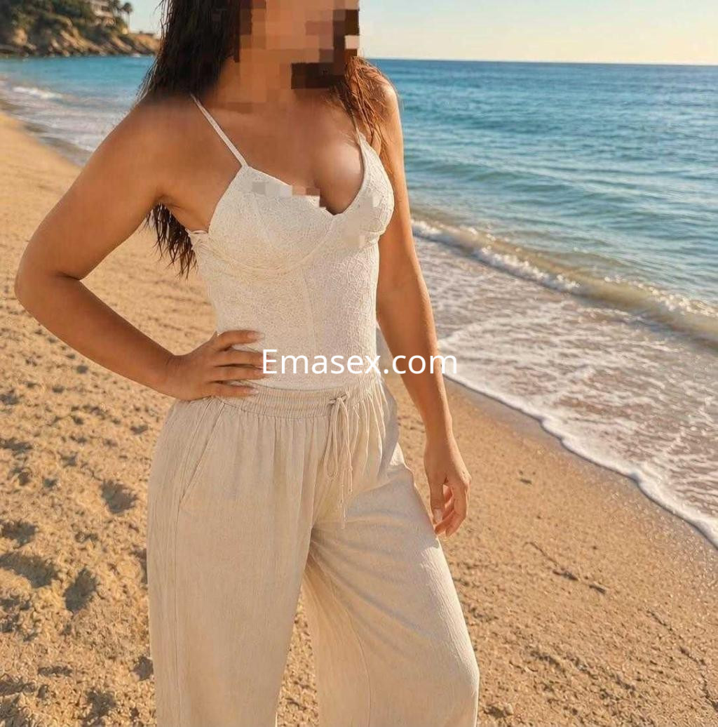 Puta en Castellón viste una blusa sin mangas blanca y pantalones claros, posando en la playa al atardecer.