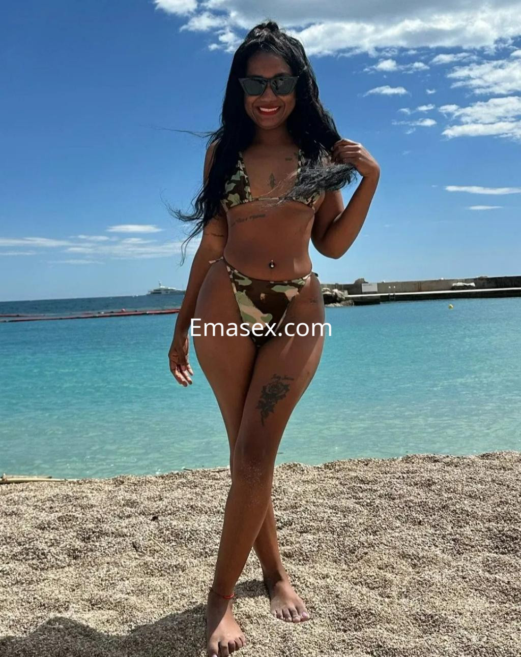 La puta en Zaragoza lleva un bikini de camuflaje y gafas de sol, posando sonriente junto al mar.