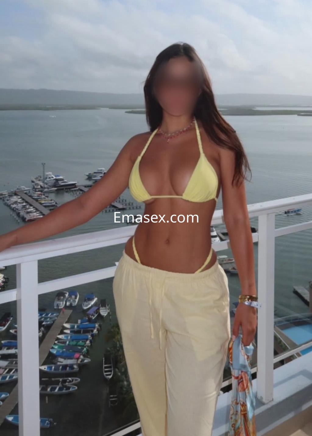 Escort en Bilbao, vistiendo un bikini amarillo y pantalones claros, posando frente a un puerto deportivo.