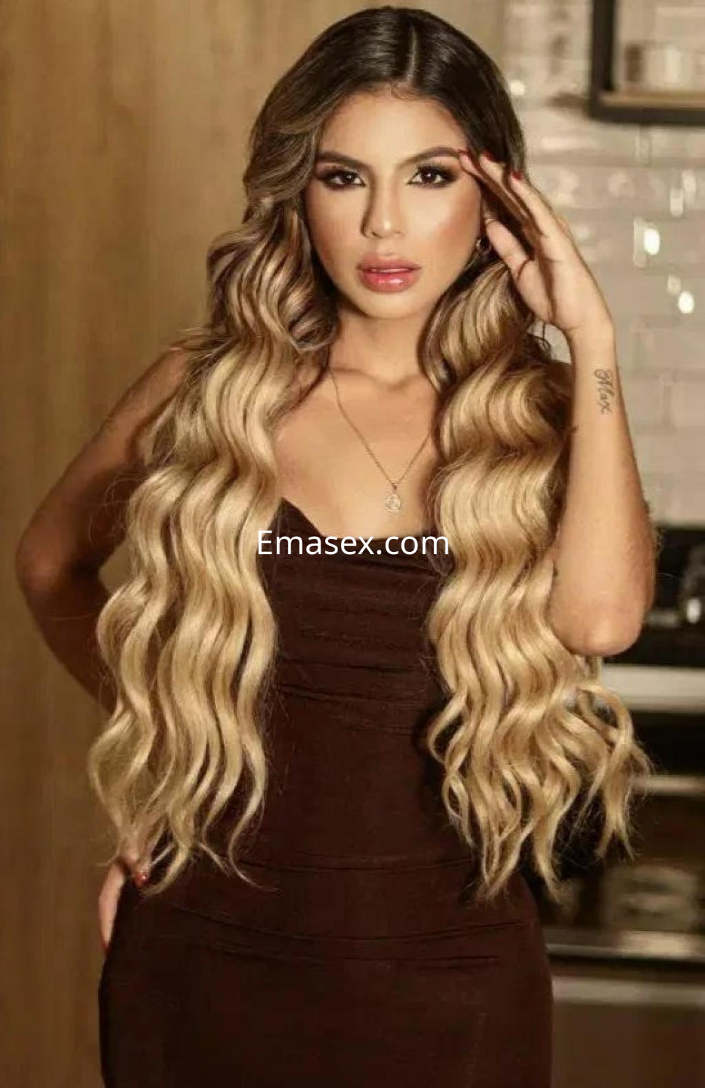 Puta en Barcelona con cabello largo y ondulado, viste un vestido ajustado marrón mientras posa con una mano en la cabeza.