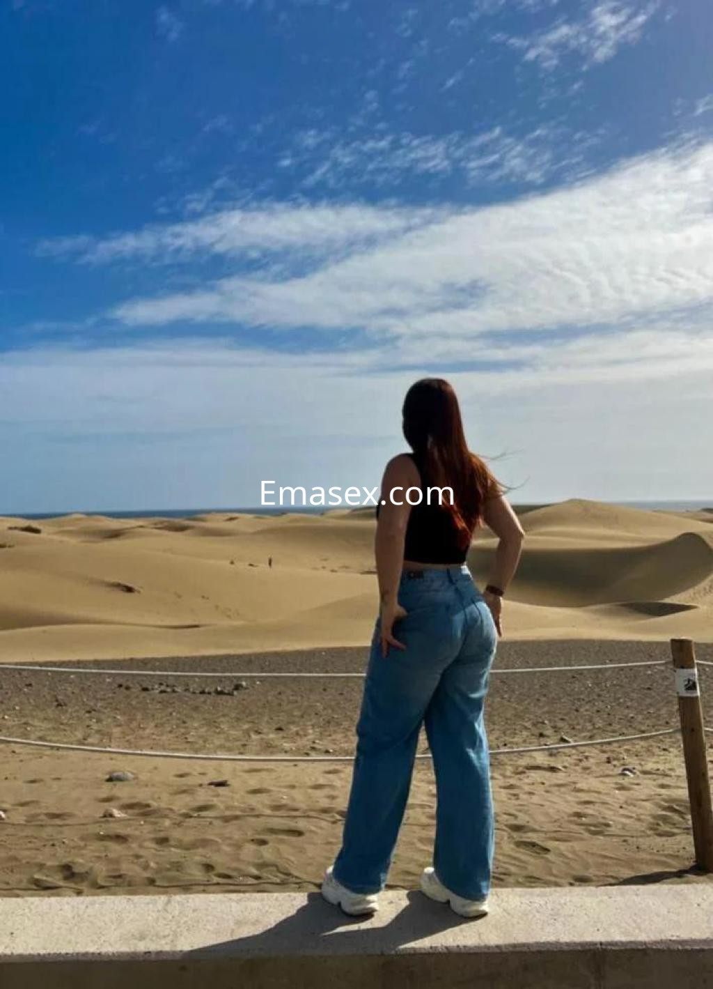 Escort en Las Palmas de espaldas, con top negro y pantalones vaqueros, frente a un paisaje de dunas.
