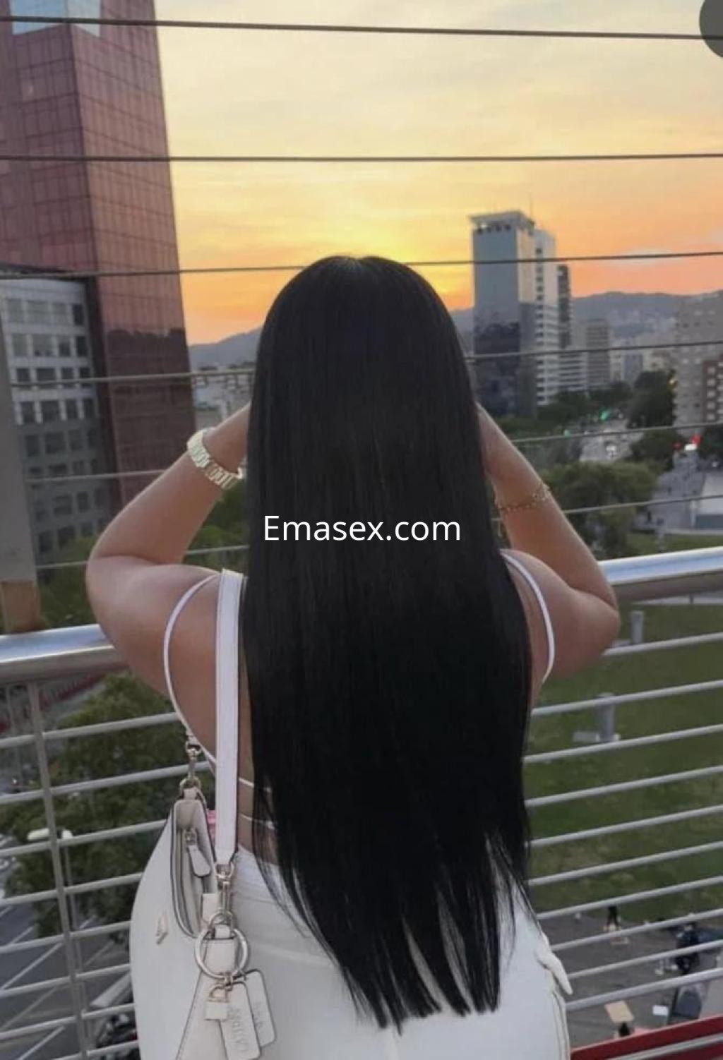 Escort en Las Palmas con cabello largo y lacio, de espaldas frente a un atardecer urbano, usando una blusa blanca.