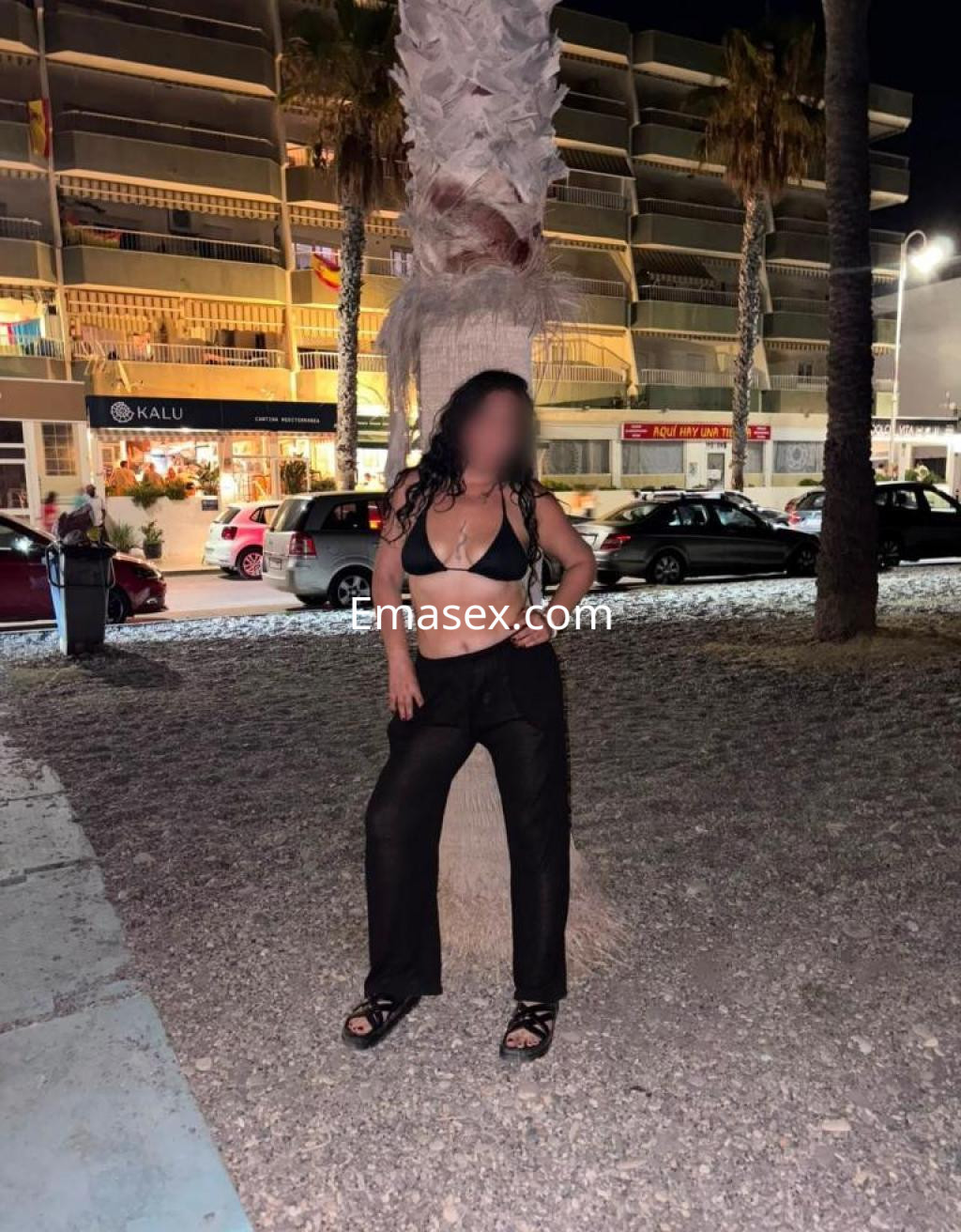 Escort en Granada, posando junto a una palmera, lleva un bikini negro y pantalones oscuros, con un fondo nocturno.