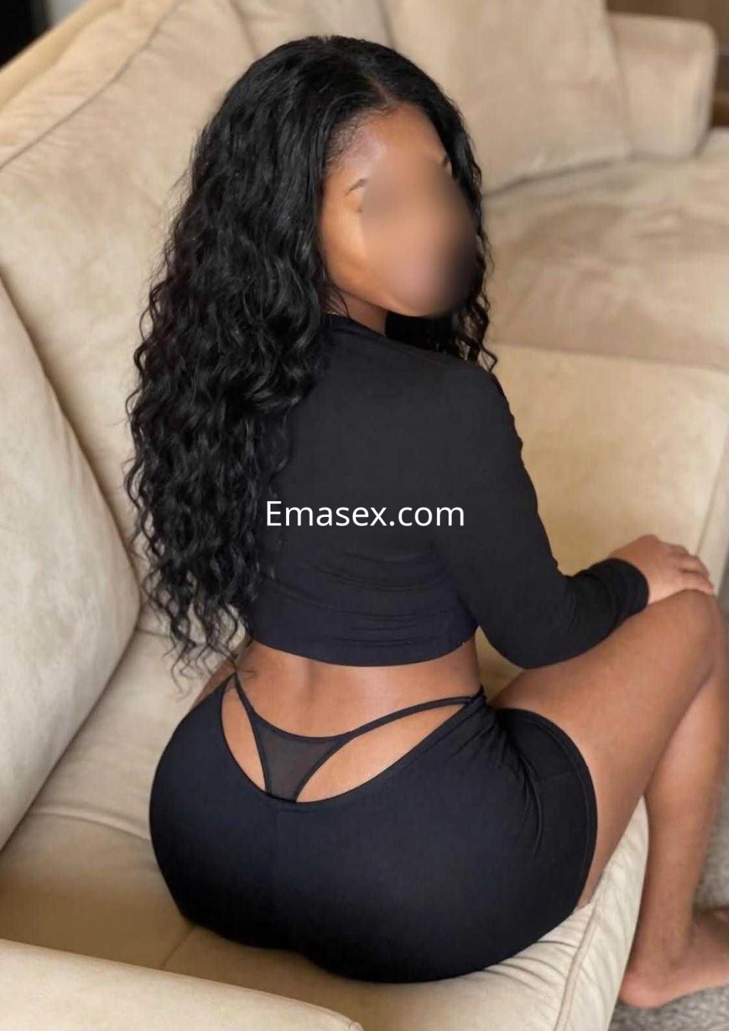 Escort en Tarragona con cabello rizado y largo, viste un conjunto negro de top y falda corta, sentada de espaldas.