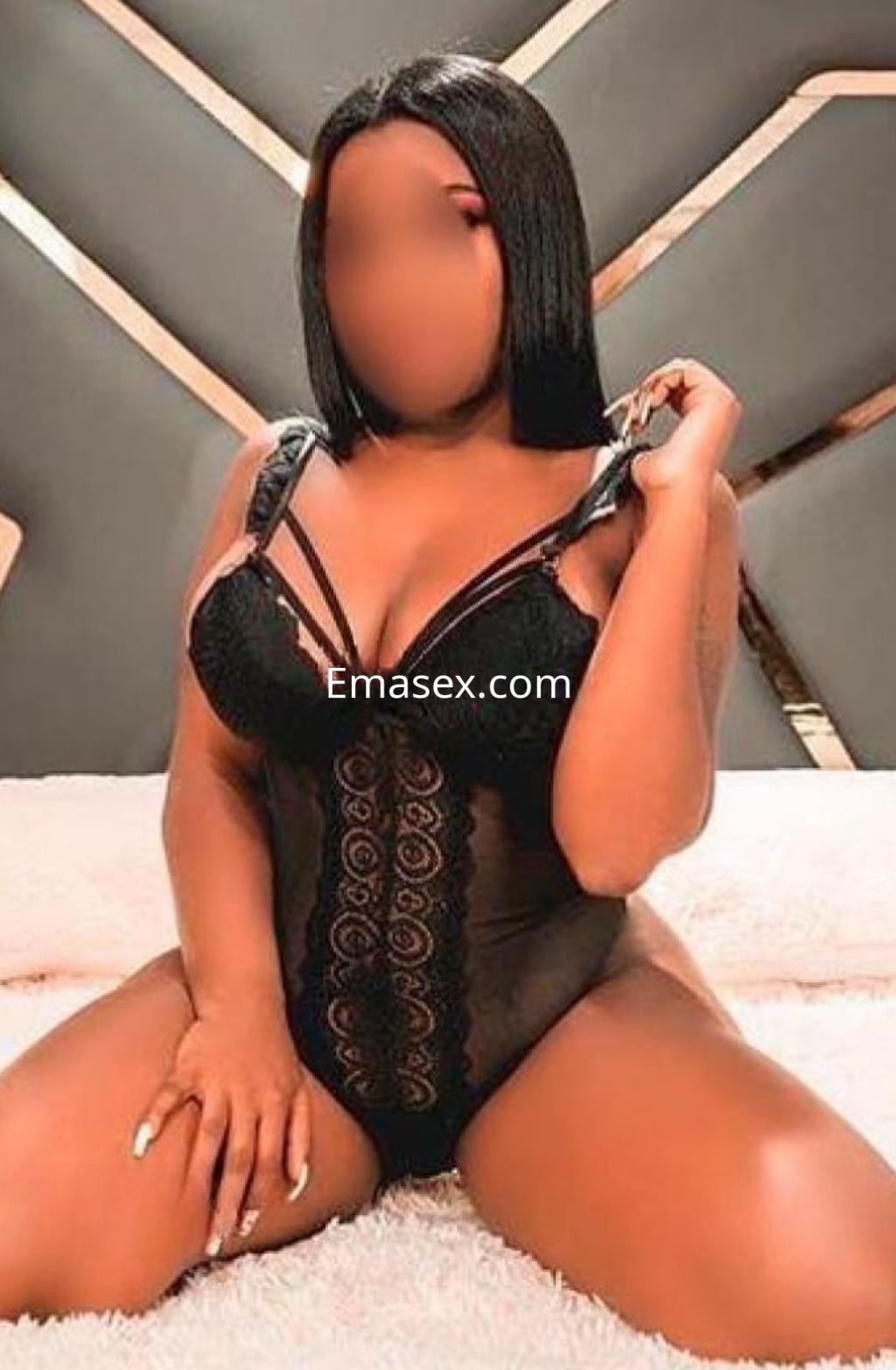 Escort en Barcelona sentada sobre una superficie clara, vistiendo un body negro con detalles de encaje y cabello lacio.
