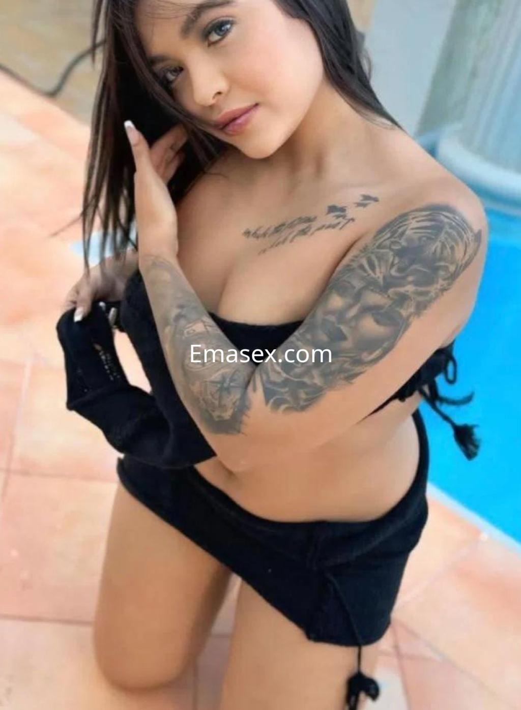 La escort se arrodilla sobre el suelo de cerámica en Almería, luciendo un bikini negro y un tatuaje en su brazo.
