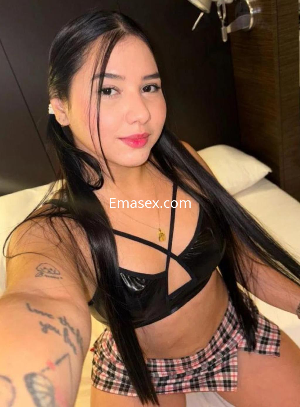 Puta en Almería posando en una habitación con una blusa negra de tirantes y falda corta de cuadros.