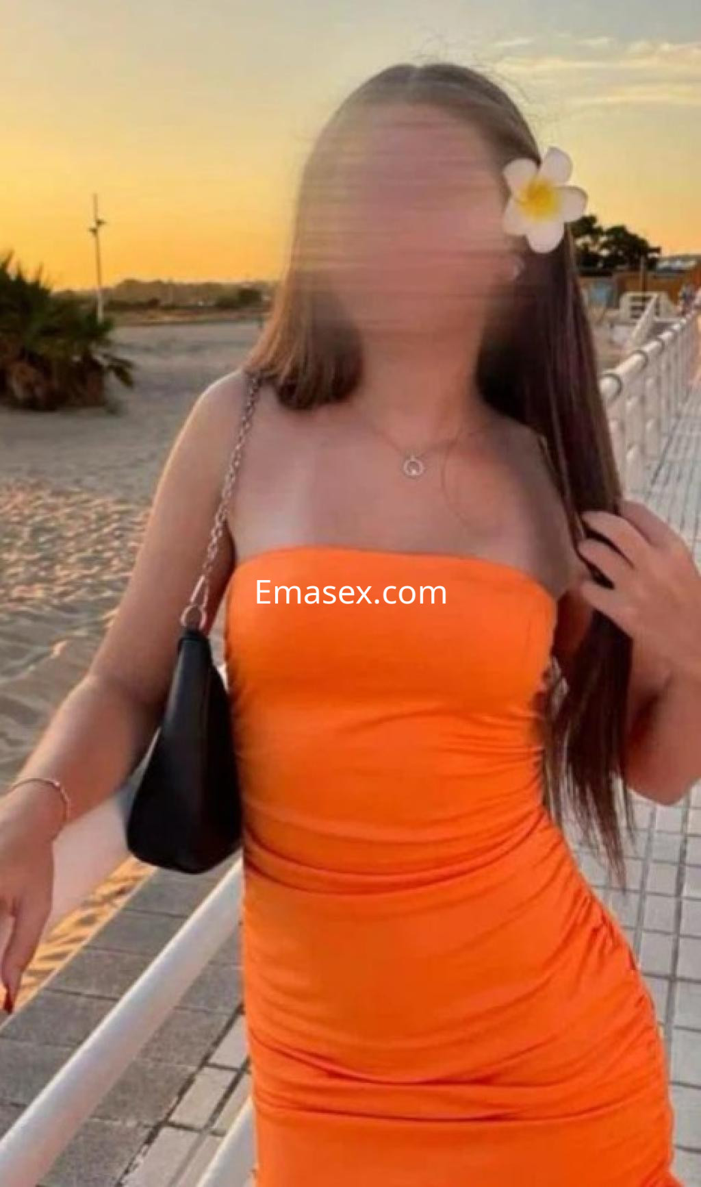 Puta en Madrid con cabello largo y lacio, vistiendo un vestido naranja ajustado y una flor en el cabello al atardecer.