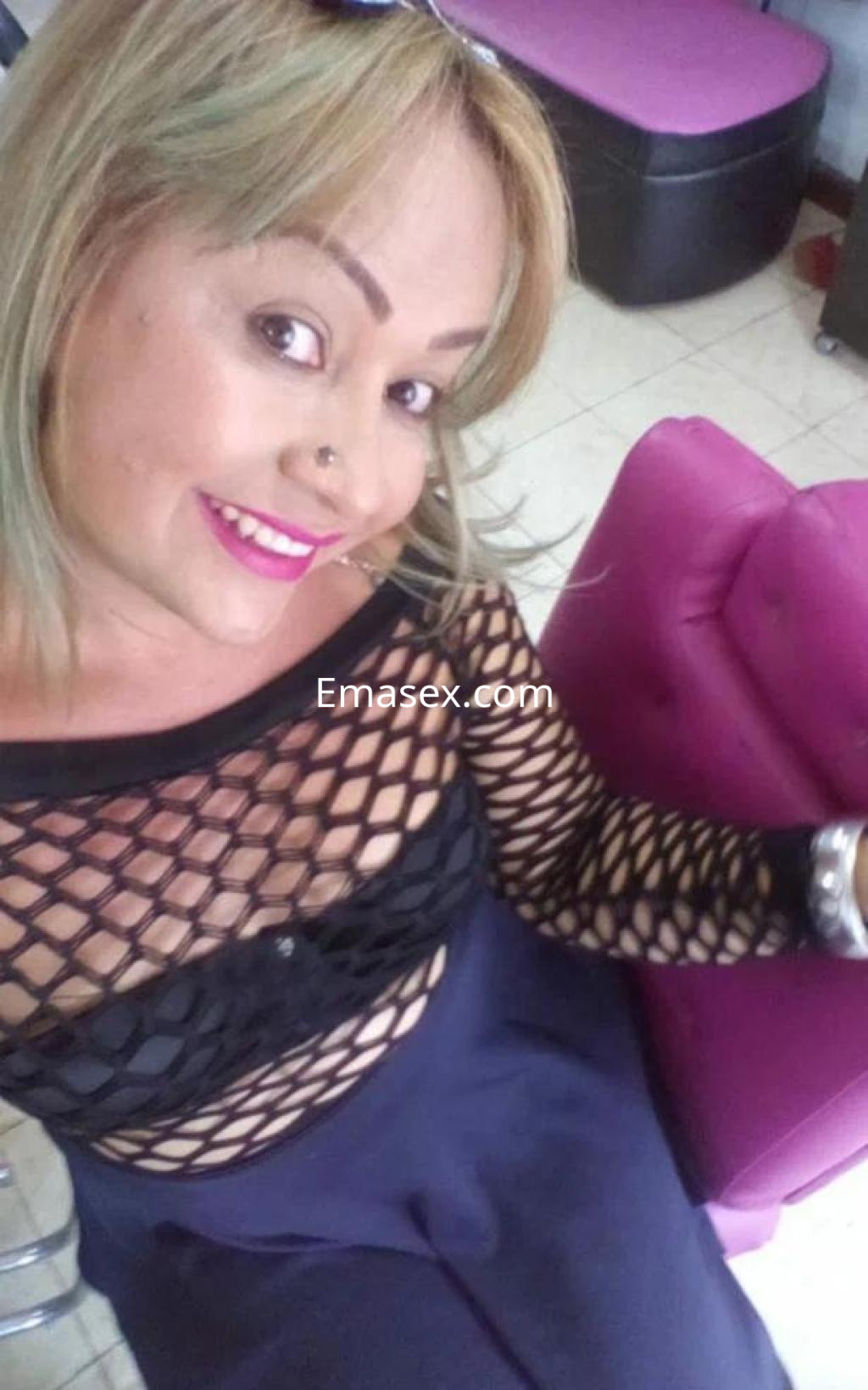 Puta en Coruña con blusa de malla negra y falda larga, posando sonriente en un ambiente interior.