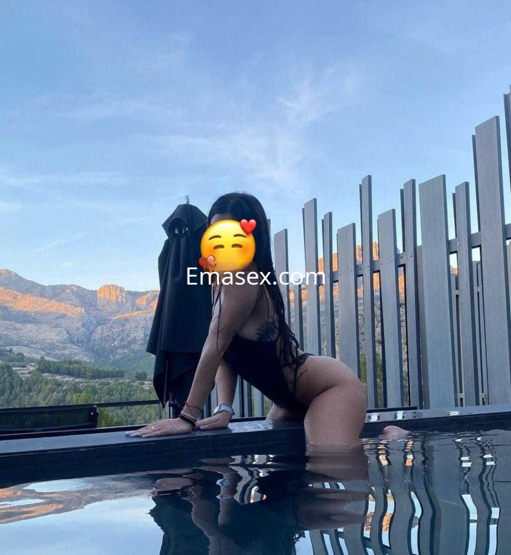 Puta en Murcia posando en una piscina al atardecer, con un traje de baño negro y un paisaje montañoso de fondo.