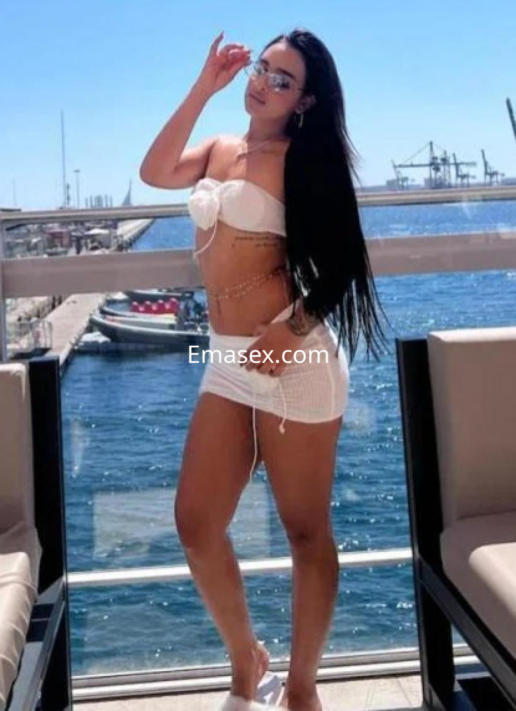 La puta en Madrid lleva un bikini blanco, pareo corto y gafas de sol, posando con el mar de fondo.