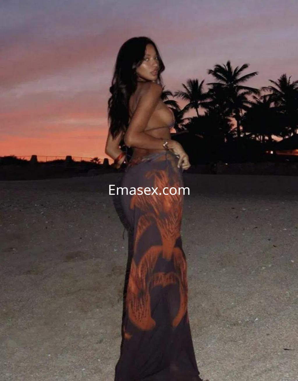 La escort en Girona se muestra de espaldas con un vestido largo y transparente, disfrutando del atardecer en la playa.