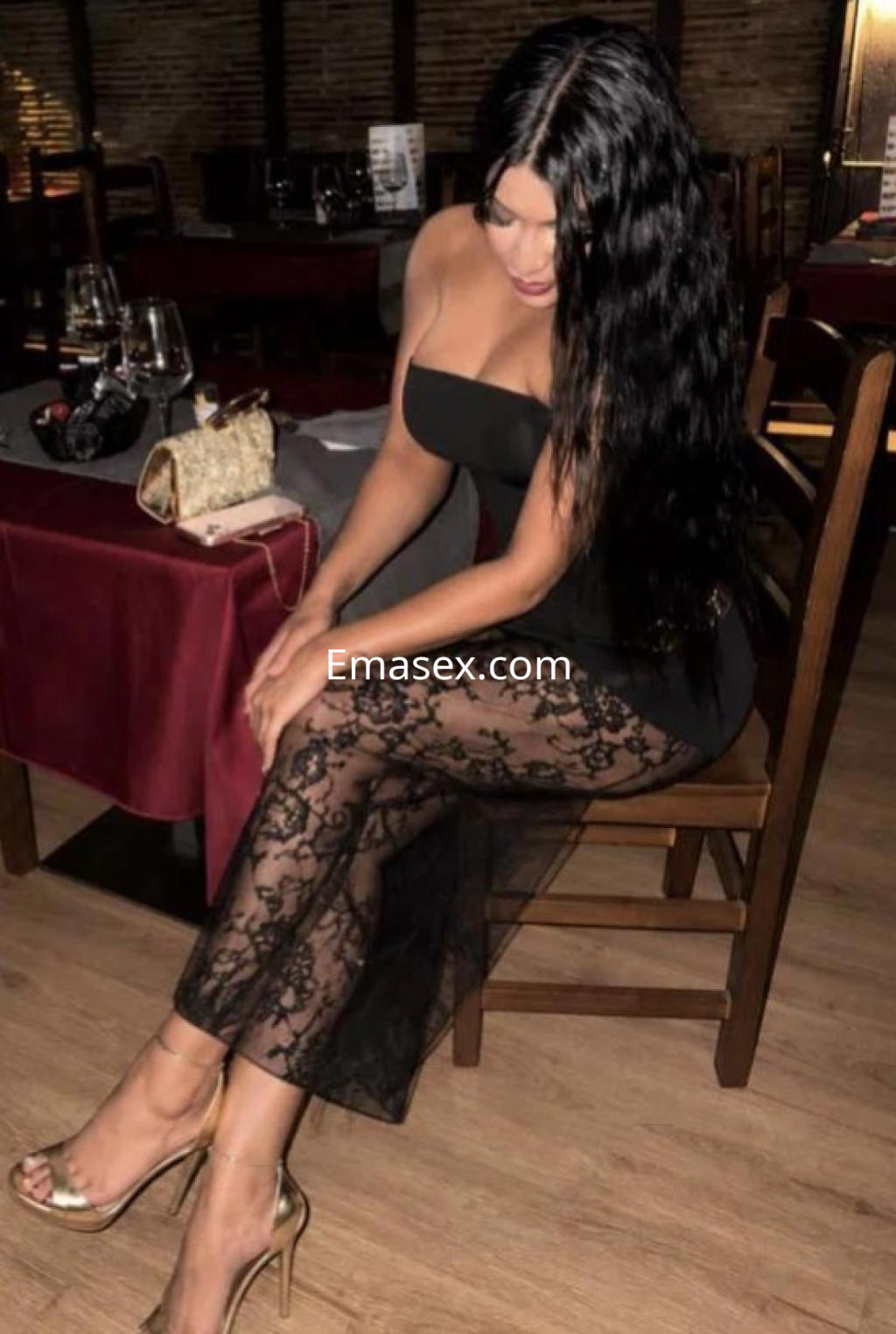 Escort en Madrid sentada en un restaurante, luce un vestido negro ajustado con encaje y tacones dorados.