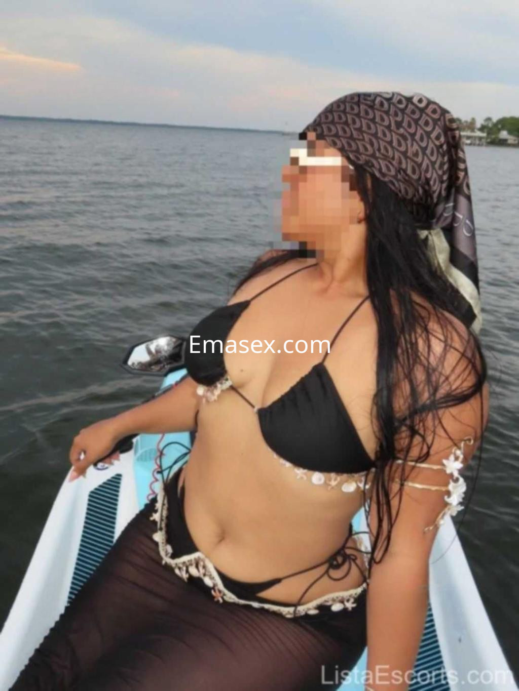 Escort en Toledo con bikini negro y accesorios de playa, sentada en una embarcación junto al agua.