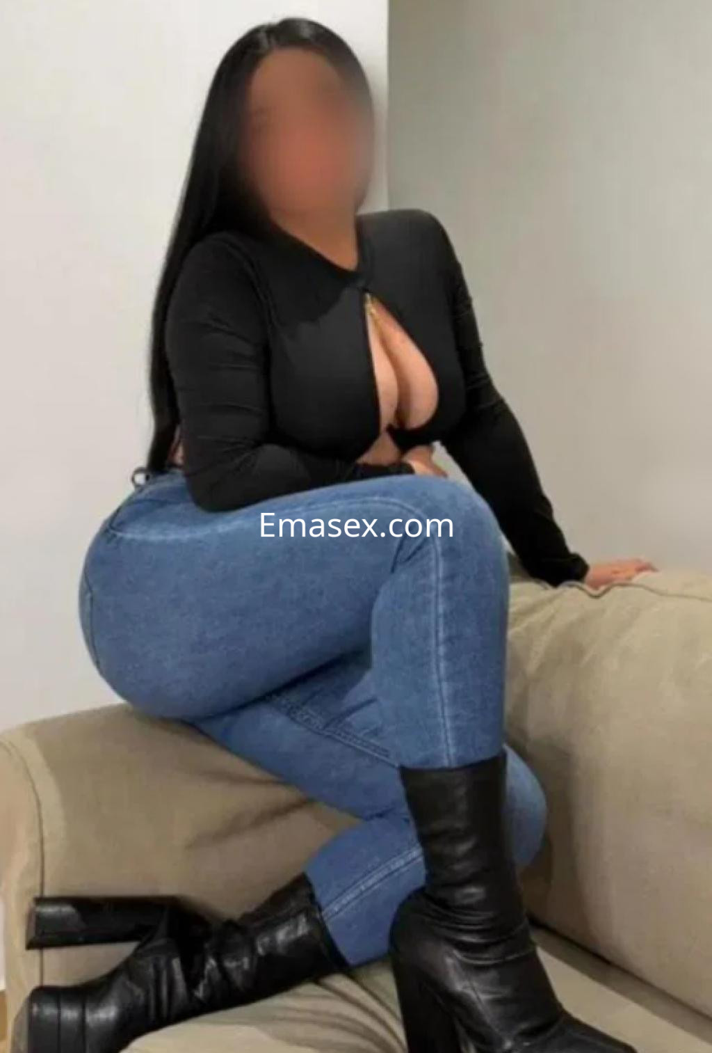 Escort en Logroño posando en un sofá, viste una blusa negra ajustada y jeans ceñidos, con botas altas.