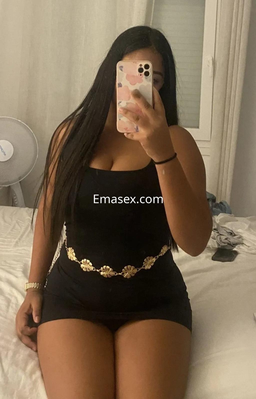 La escort en Málaga posa sentada en la cama, con un vestido negro ajustado y un cinturón decorativo dorado.