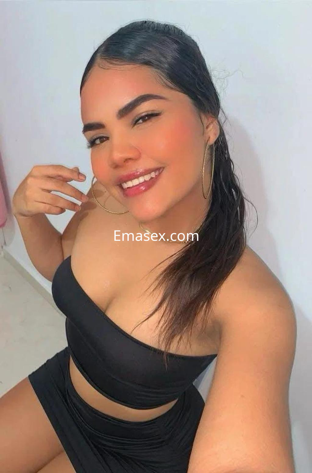 Escort en Sevilla con cabello oscuro y lacio, vistiendo un conjunto negro que realza su figura, sonriente en un fondo claro.