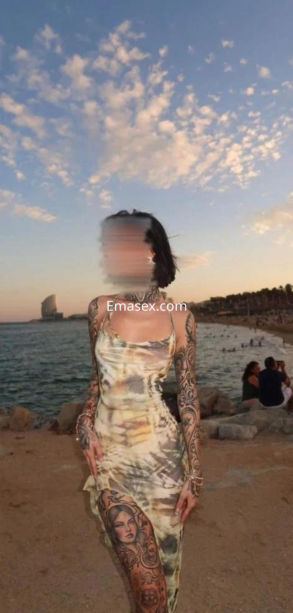 Puta con un vestido transparente y estampado, mostrando sus tatuajes, posando frente al mar en Sevilla.