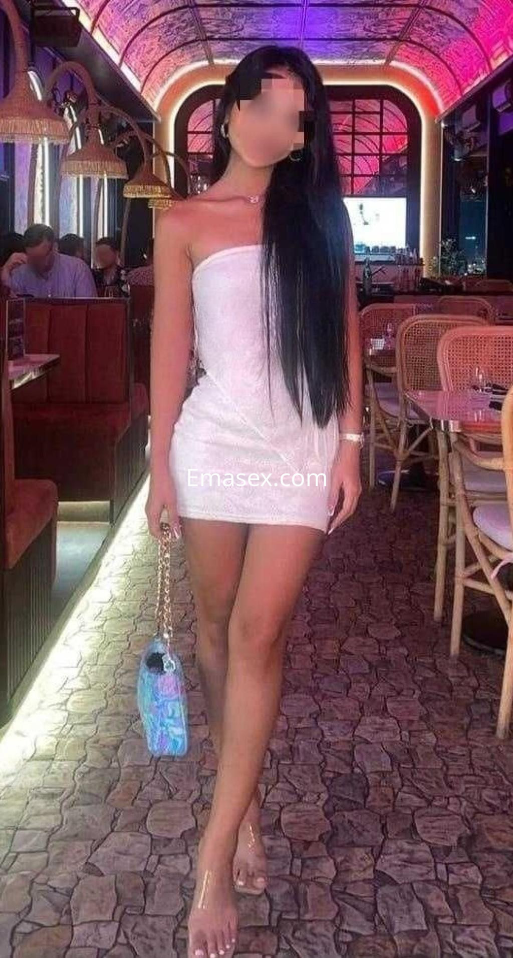 Escort en Alicante con vestido blanco ajustado y cabello largo, posando en un restaurante elegante.