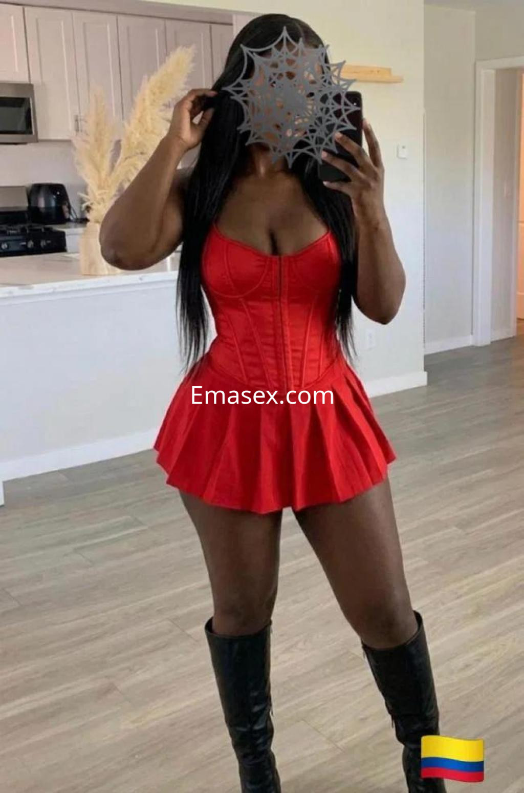 Puta en Cáceres con un vestido rojo ajustado y botas altas, posando frente a un espejo en un ambiente moderno.