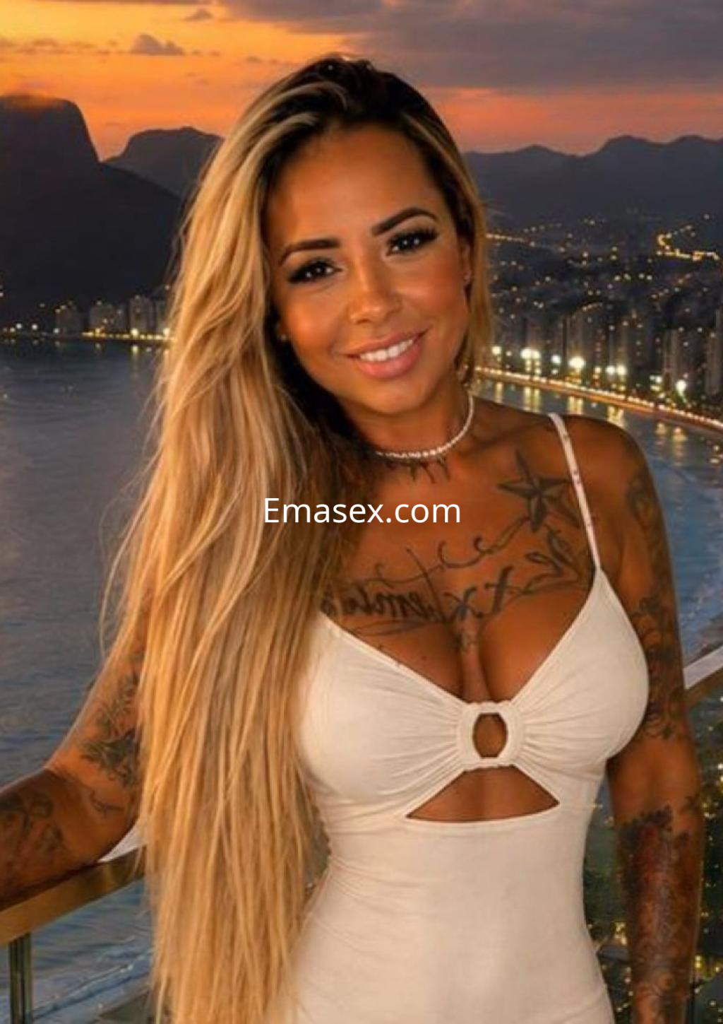 Puta con cabello largo y rubio, vestido blanco con corte en el pecho, sonríe frente a un atardecer en Alicante.