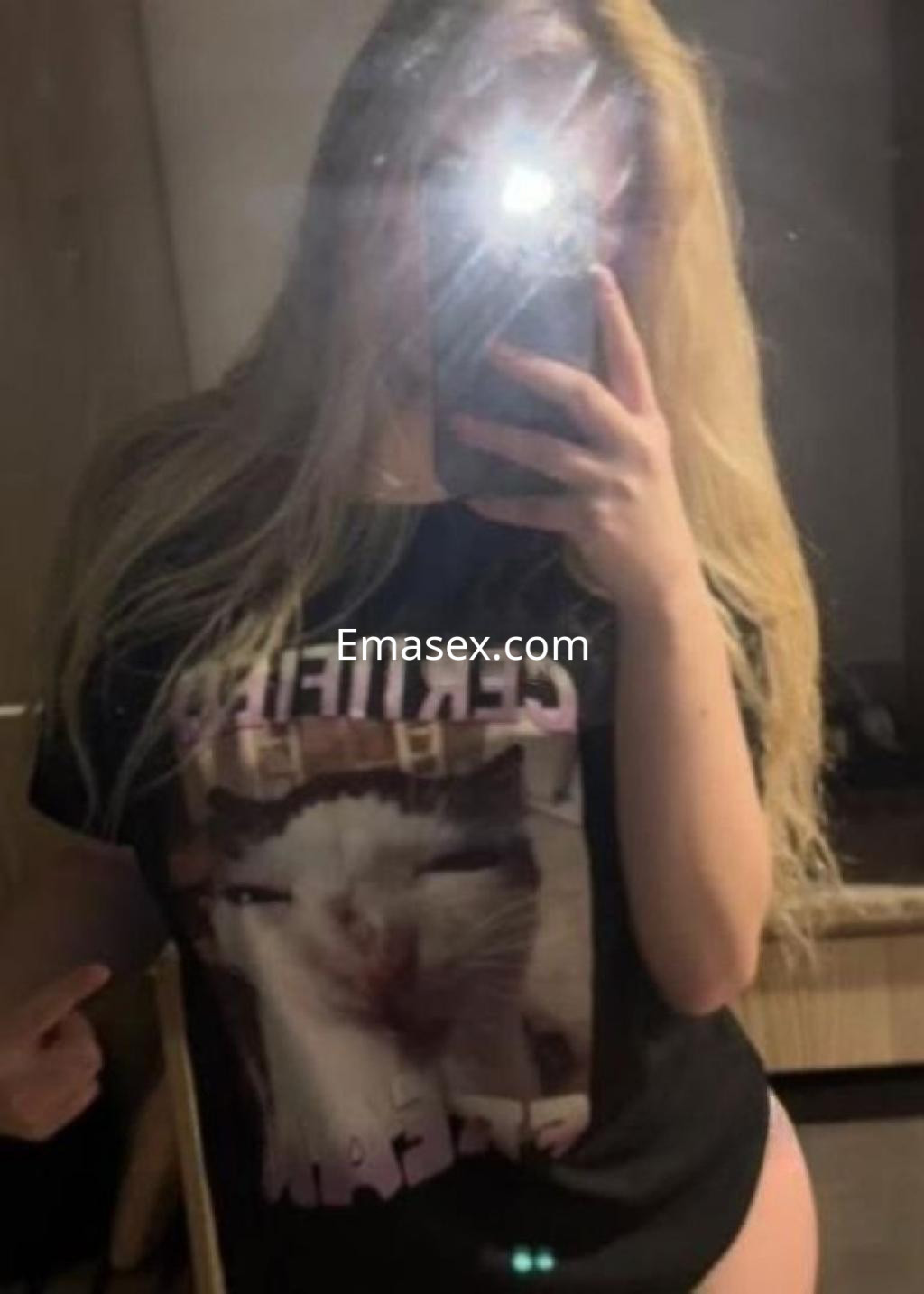 Puta en Castellón posa frente al espejo con una camiseta negra de un gato y el pelo rubio suelto.