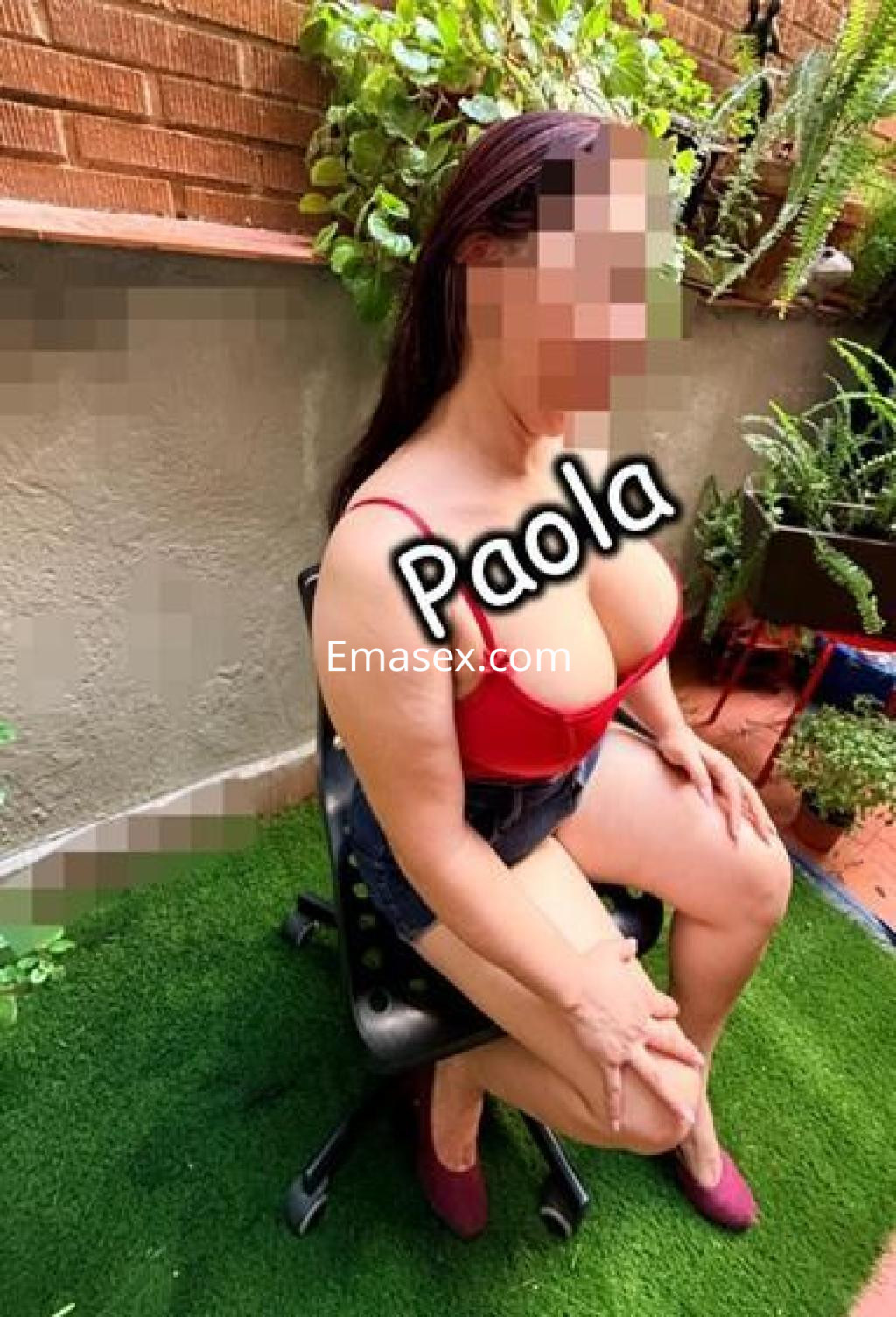 Escort en Madrid sentada en una silla, luciendo un top rojo y pantalones cortos, rodeada de plantas verdes.