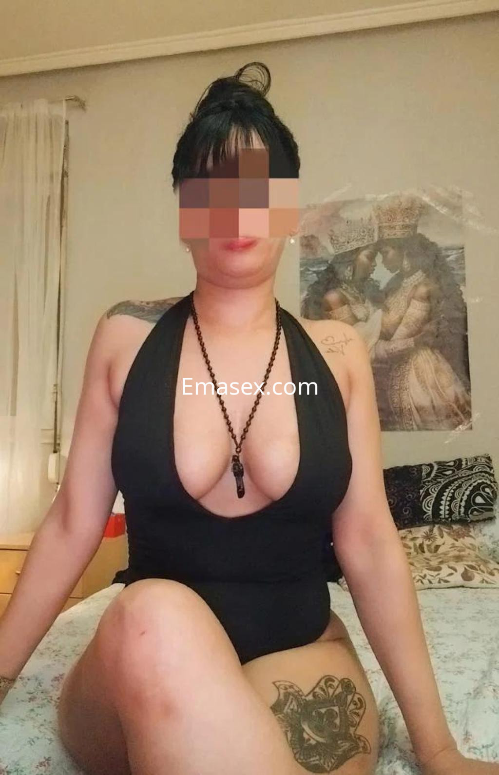 Puta en Oviedo con un traje de baño negro, sentada en una cama con tatuajes visibles y un peinado recogido.