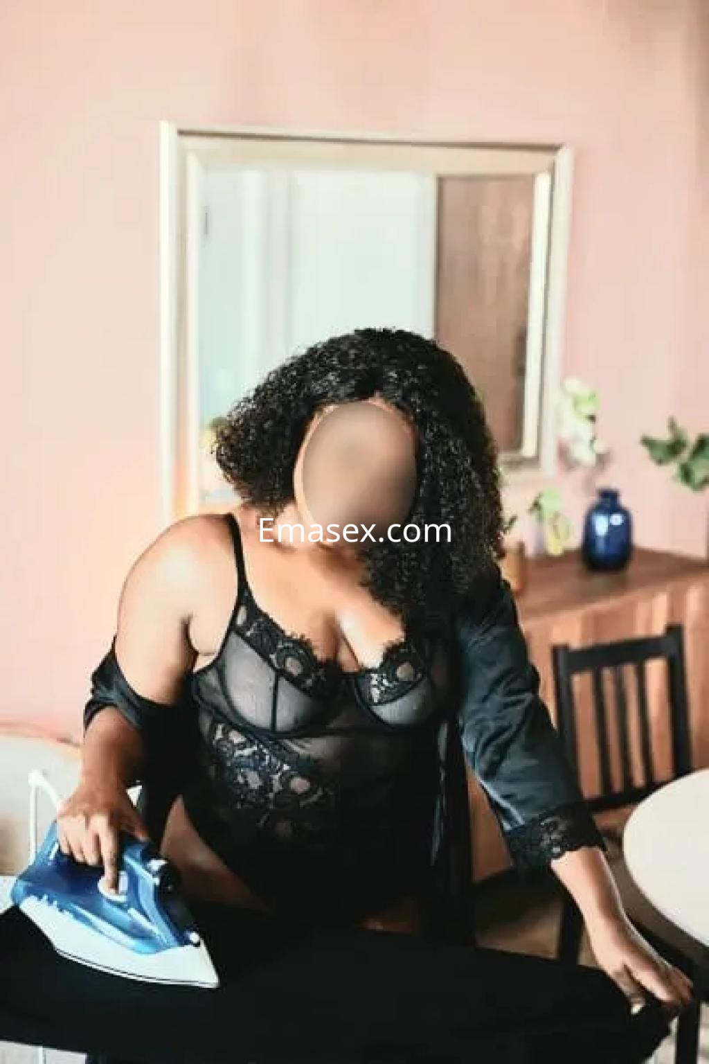 Escort en Oviedo viste lencería negra y se encuentra frente a una mesa planchando, con cabello rizado y un fondo decorativo.