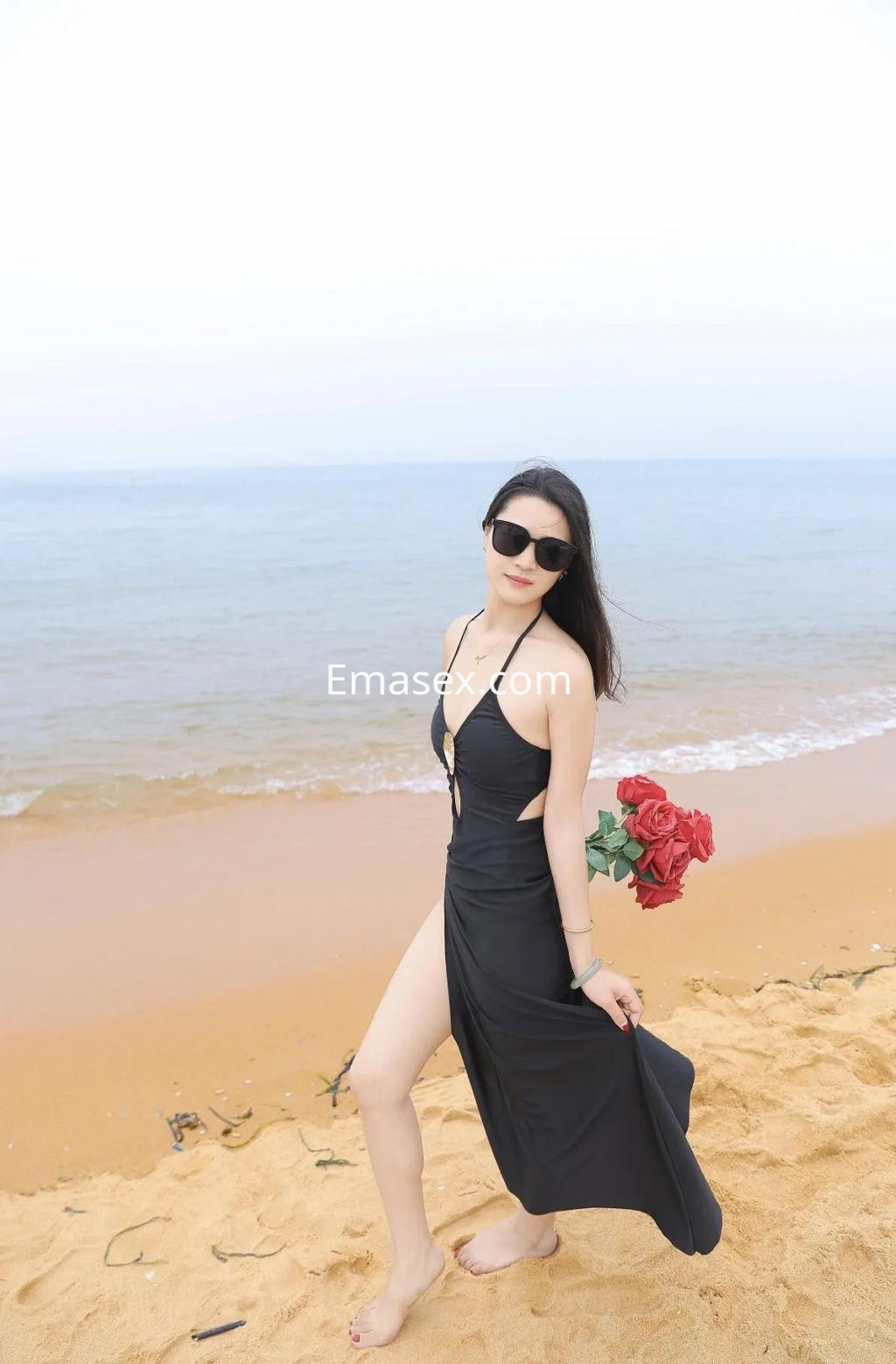 Escort en Murcia con un vestido negro, sandalias, gafas de sol y un ramo de rosas en la playa.