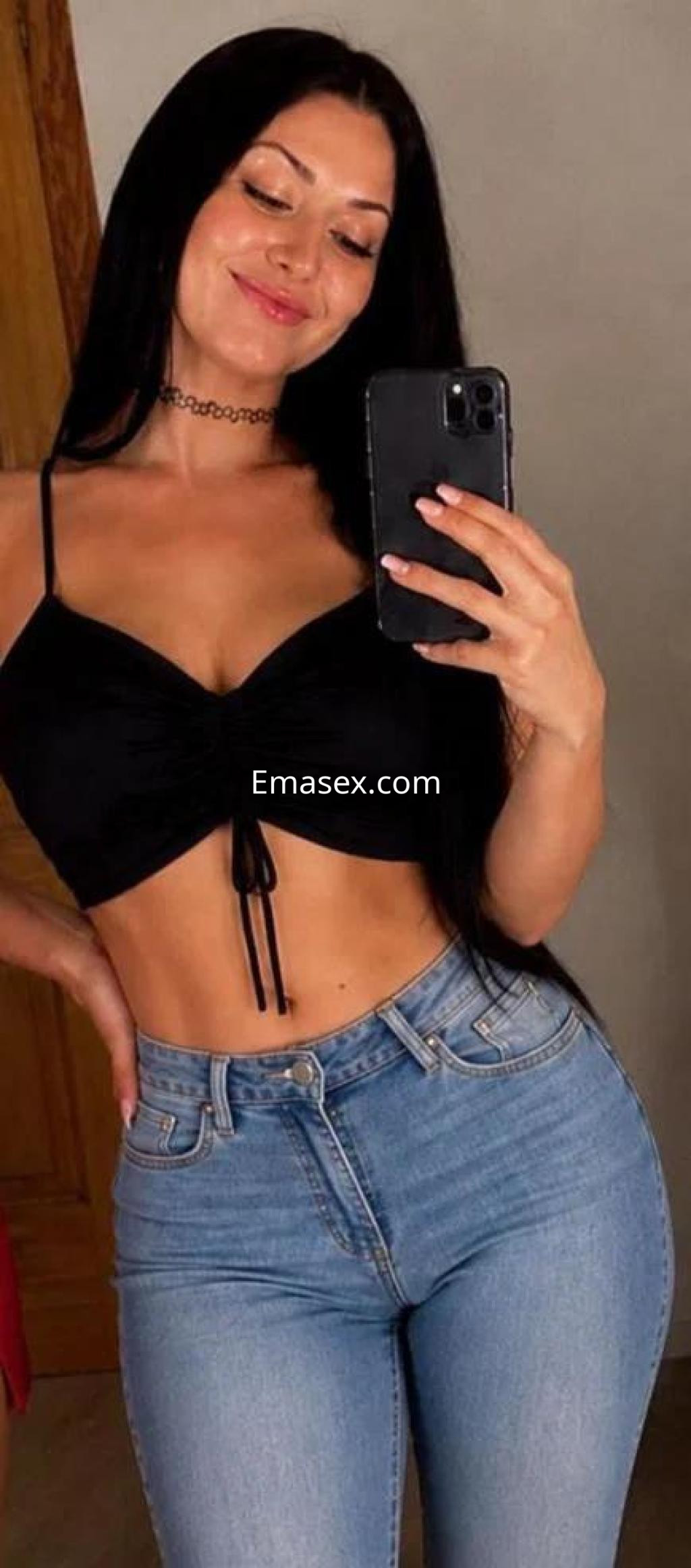 Escort en Murcia posando con una blusa negra ajustada y jeans claros, mostrando una sonrisa mientras se toma una selfie.
