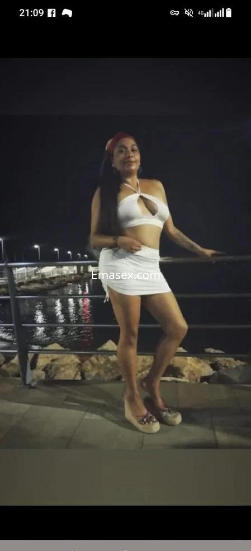 Escort en Murcia, posando con un vestido blanco ajustado y tacones, junto a un muelle iluminado por la noche.