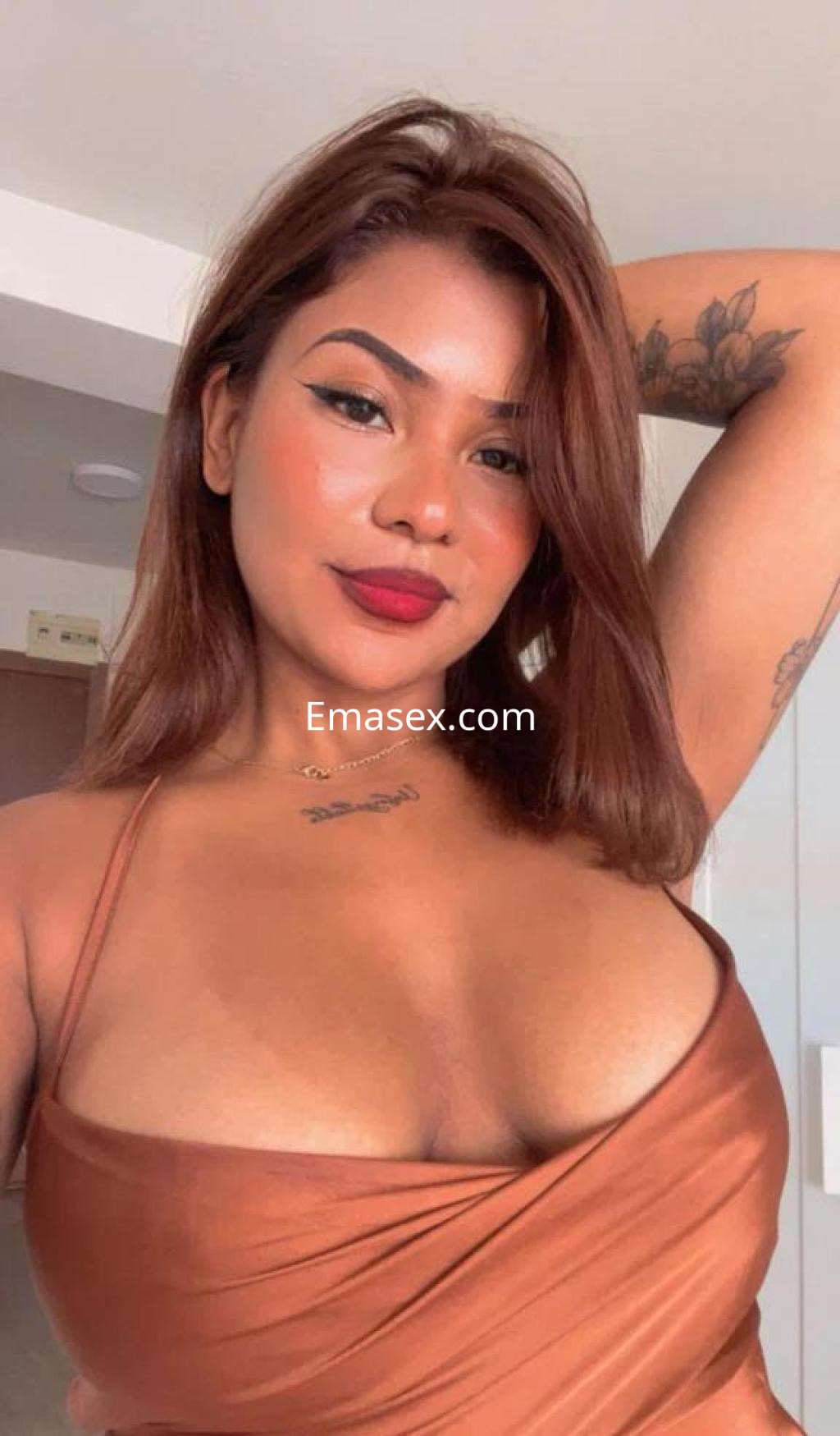 Escort en Alicante posando con una blusa marrón que resalta su figura, mostrando su cabello castaño y tatuajes.