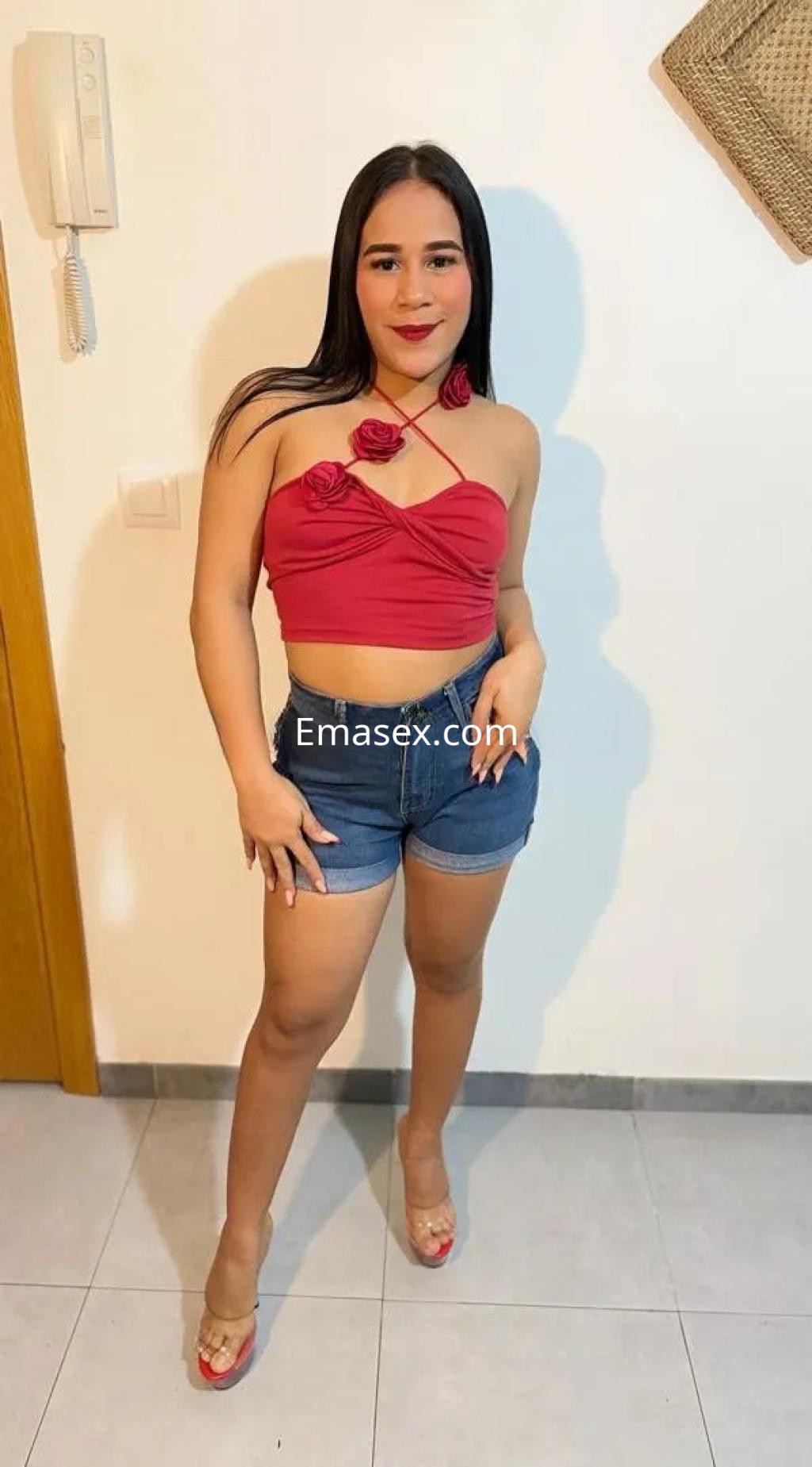 Escort en Córdoba con blusa roja con flores y shorts de mezclilla, posando con una sonrisa en un ambiente interior.