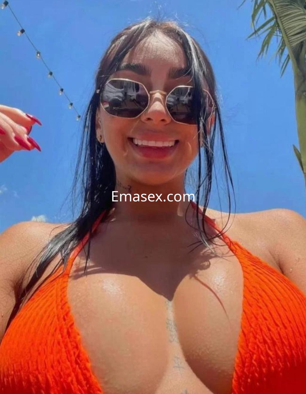 La escort sonríe a la cámara en Madrid, luciendo un bikini naranja y gafas de sol bajo un cielo azul.