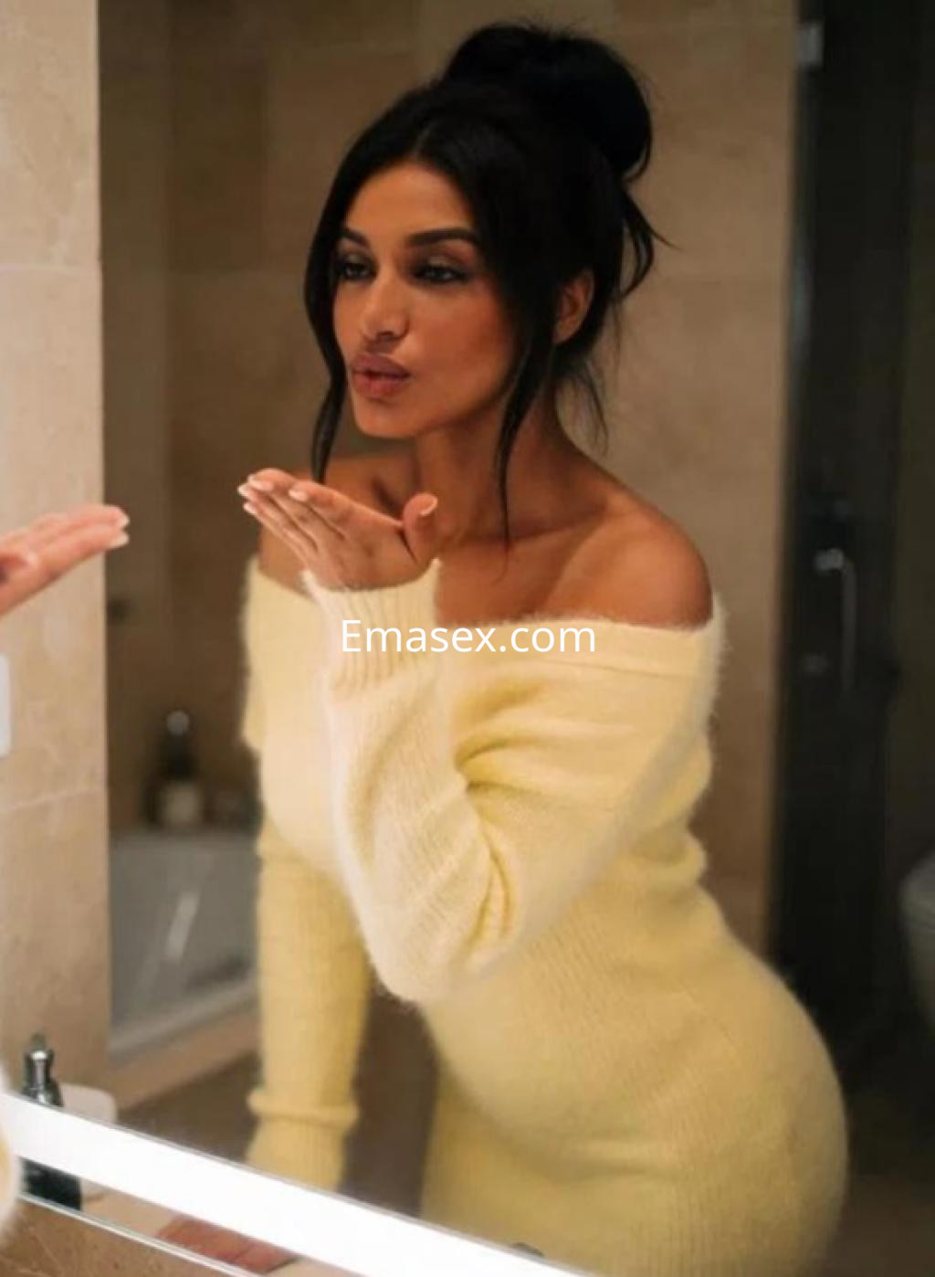 Escort con cabello oscuro recogido, viste un suéter amarillo de manga larga mientras se mira en el espejo en Madrid.