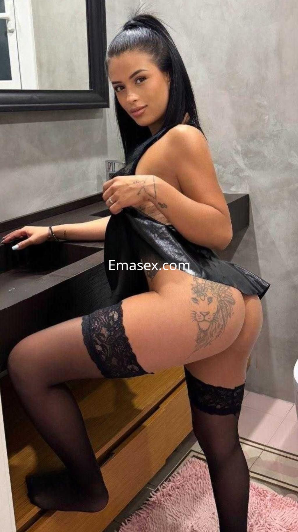 Puta en Barcelona de pie junto a un tocador, usando un corset negro y medias con encaje, mostrando un tatuaje en su muslo.