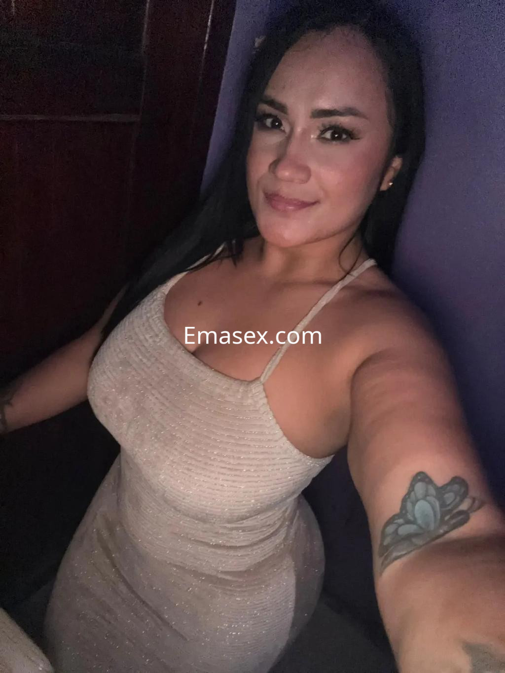 Escort en Barcelona con vestido ajustado y brillante, cabello largo y lacio, sonríe en un ambiente íntimo.