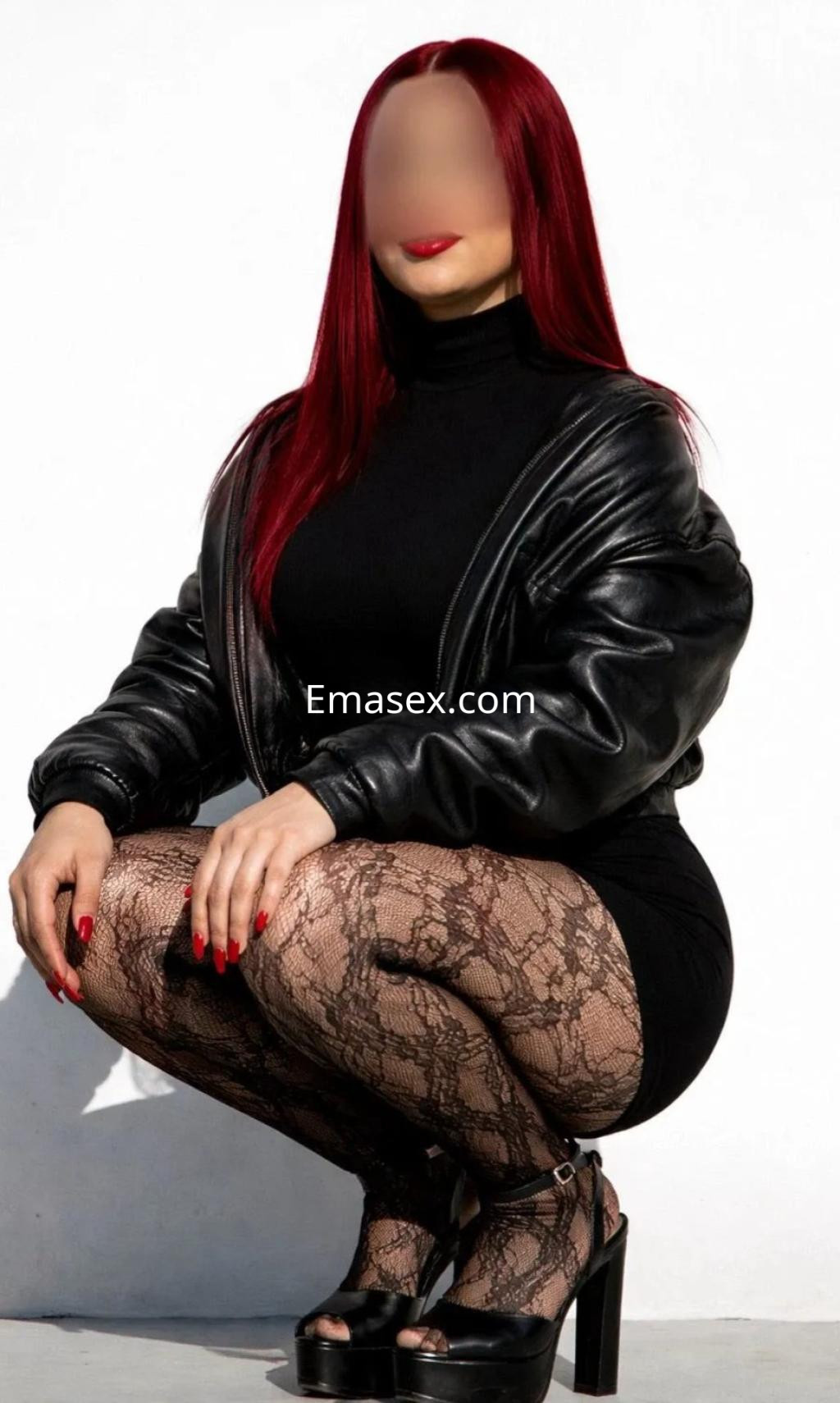 Escort en Vigo con cabello rojo, viste un vestido negro y mallas de encaje, posando en cuclillas con tacones.