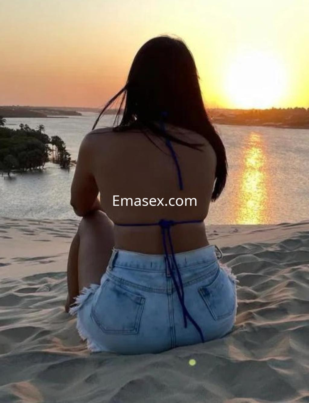 Escort sentada en una duna con vista al atardecer, vistiendo un bikini y shorts de mezclilla en Zaragoza.