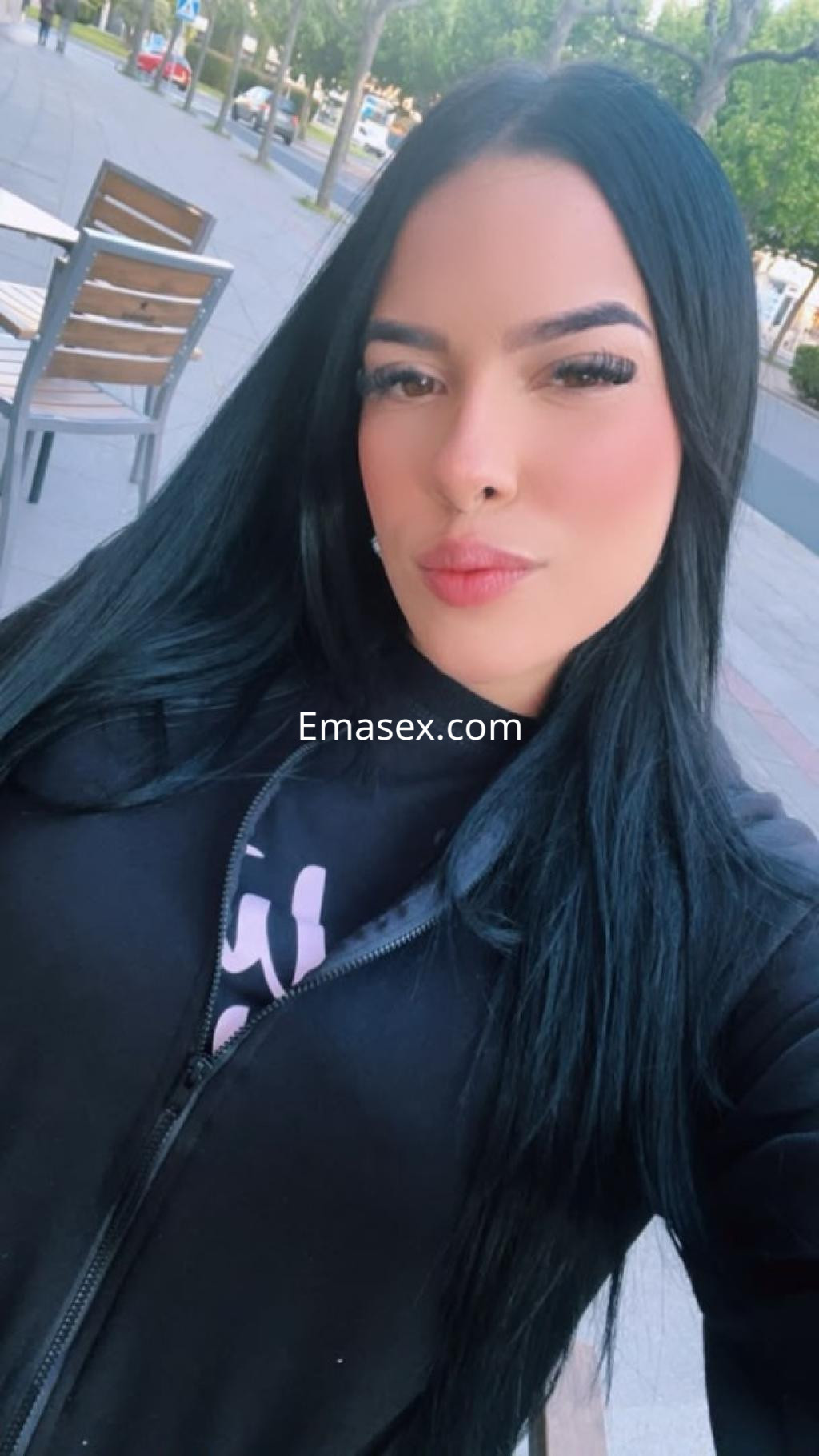 Escort con cabello largo y negro, usando una chaqueta negra, posando sonriente en una plaza en Santiago.