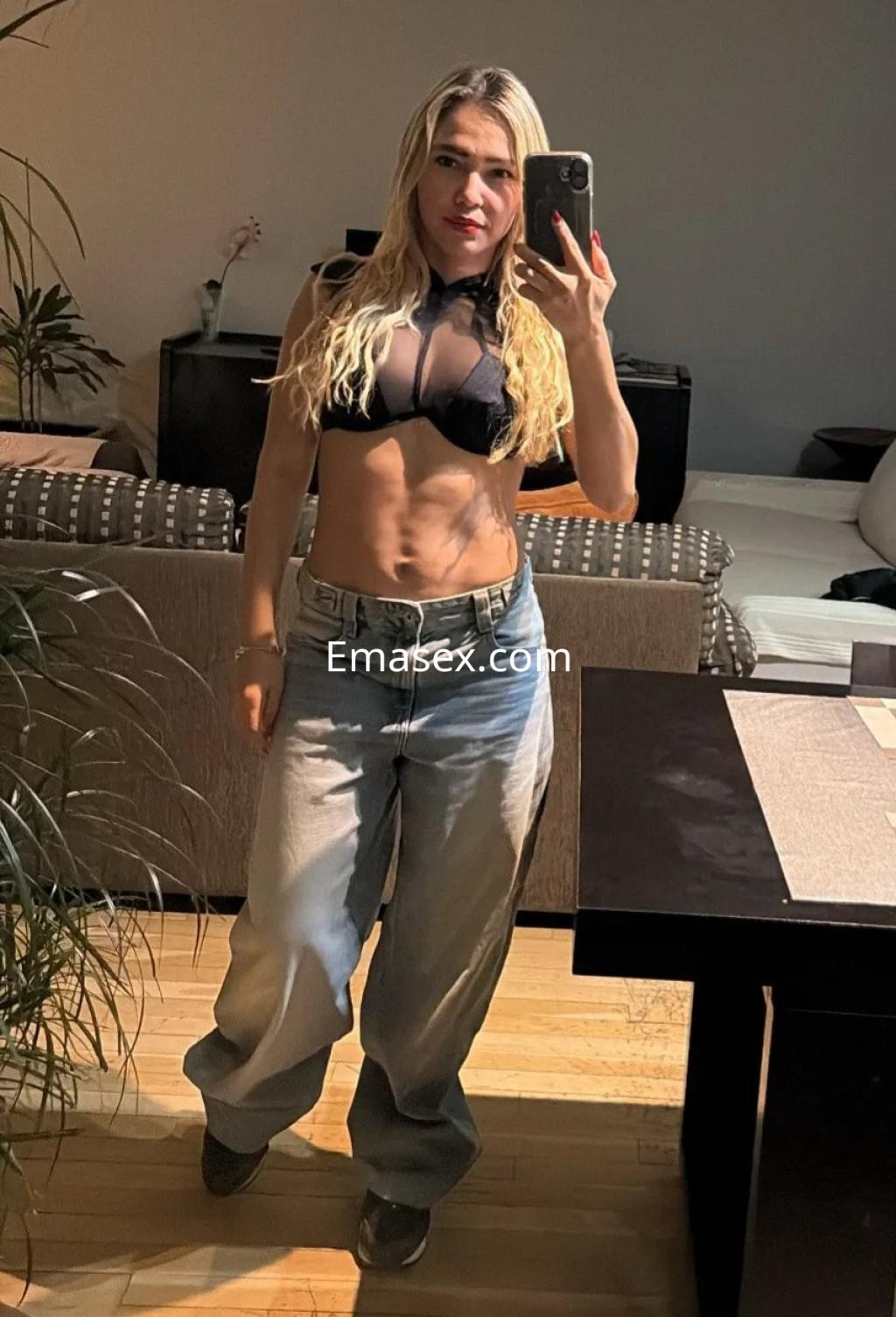 Escort en Zaragoza, posando en un interior con un top negro transparente y pantalones anchos de mezclilla.
