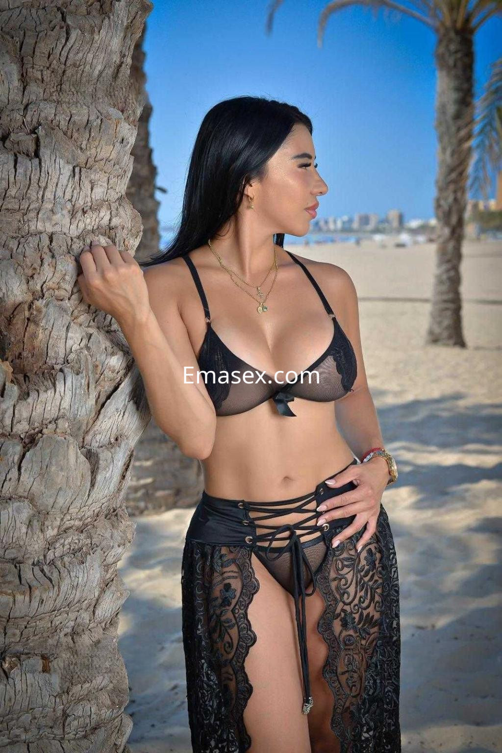 Escort en Málaga posando junto a una palmera, con un bikini negro y una falda de encaje transparente.