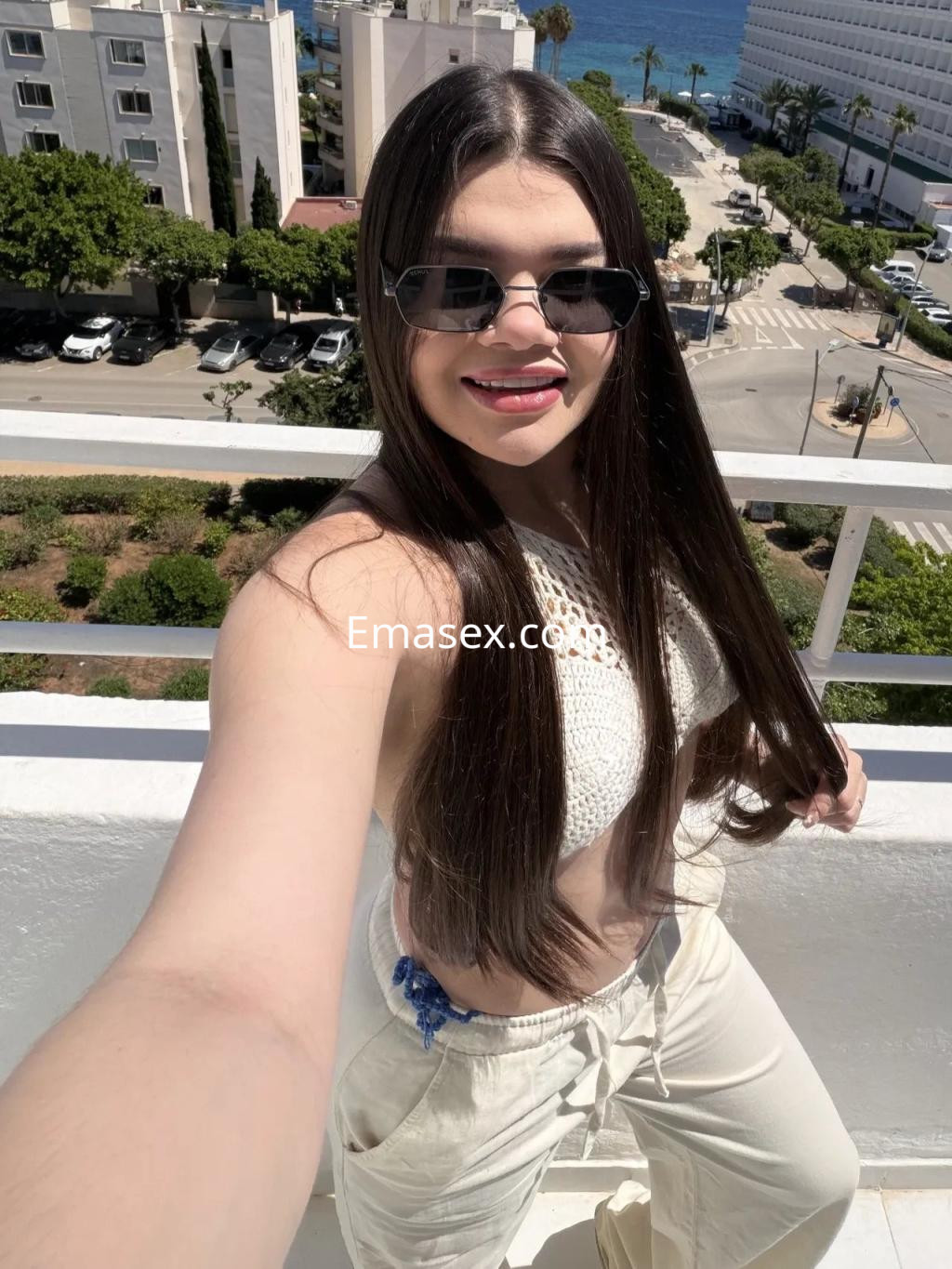 Puta posa en una terraza con vista al mar, vistiendo un top blanco y pantalones claros, con gafas de sol.
