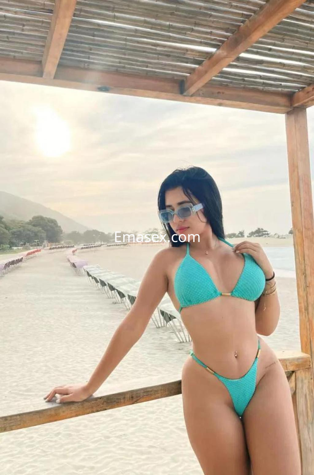 Escort en Almería luciendo un bikini azul en la playa, con gafas de sol y una pose relajada junto al mar.